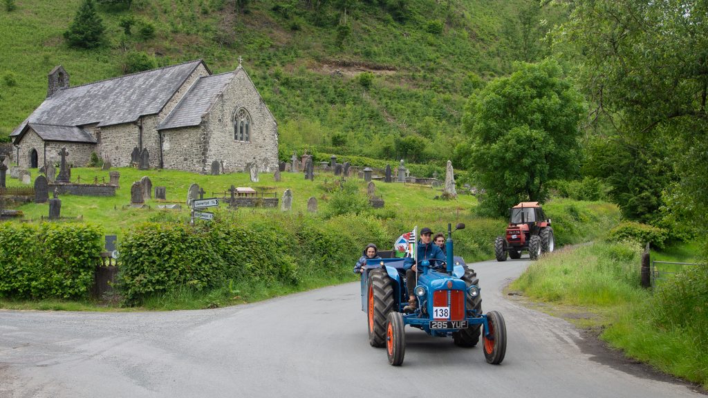 Tractor with passengers driving past St David's 11th century church at Llanwrtyd